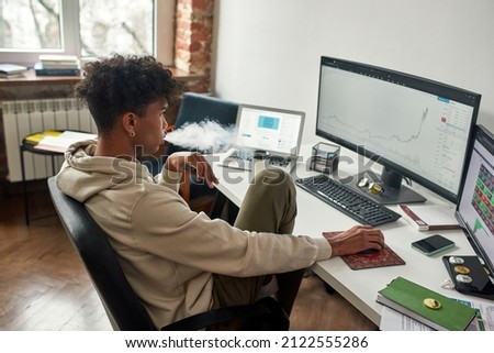 Absent-minded african american broker vaping while watching data index chart stock market. Worried young trader working with multiple screens. Working from home concept.