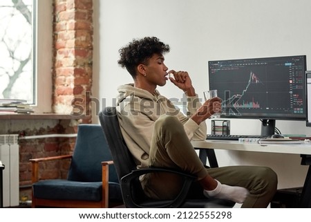 Multiethnic stock trader with glass of water taking capsule of dietary supplements for best mental work. African american trader with glass of water drinks pill while working at home modern office. Royalty-Free Stock Photo #2122555298