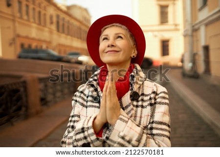 Beautiful charming blonde woman of 50s putting palms in prayer position, looking up standing on street in her favorite city, being thankful for opportunity to travel again, wearing red hat