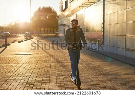 Man using electric scooter outdoor in the sidewalk. Bearded Man ready to discover the urban city at sunset with escooter. Royalty-Free Stock Photo #2122601678