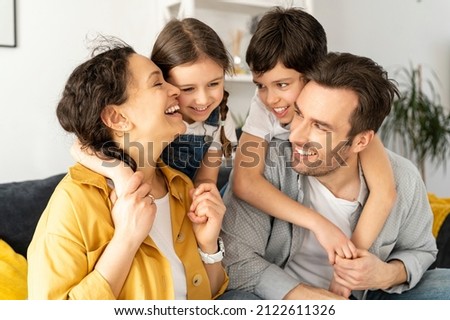 Happy together. Portrait of cheerful multiracial family of four - mom, dad, son and toddler daughter sitting on the sofa in living room, looking to each other and laughing, enjoying weekend at home Royalty-Free Stock Photo #2122611326