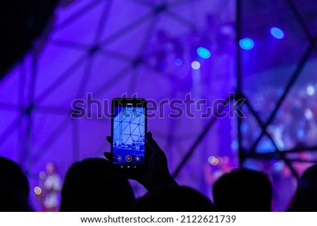 The raised hand of a person with a smartphone in front of a bright light is recording a night show. Unrecognizable people take pictures and videos on the phone in front of the show stage.