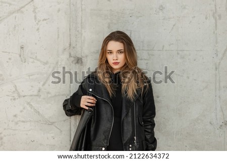 Fashionable adorable woman model with fashion black leather jacket and black hoodie with leather bag stands near a gray concrete wall Royalty-Free Stock Photo #2122634372