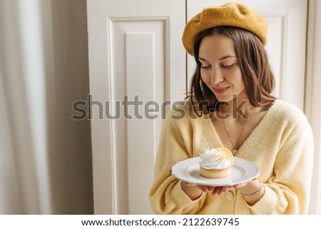 Close-up of cute caucasian young girl modestly holding plate of cake in her hands on white background. Dark-haired beauty in yellow headdress looked down. Good morning concept