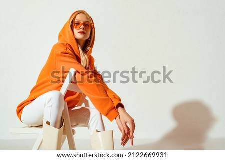 Young confident blonde girl wearing trendy orange hoodie, color sunglasses, posing on white background. Studio fashion portrait. Copy, empty space for text Royalty-Free Stock Photo #2122669931