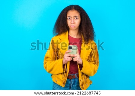 Portrait of a confused beautiful teenager girl wearing yellow jacket over blue background holding mobile phone and shrugging shoulders and frowning face.