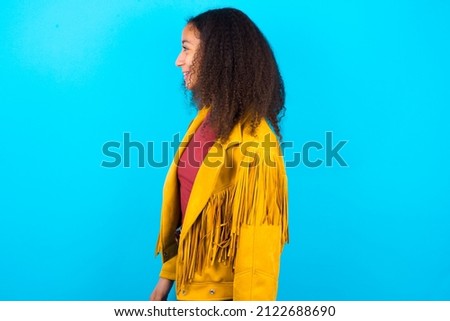 Profile of smiling beautiful teenager girl wearing yellow jacket over blue background with healthy skin, has contemplative expression, ready to have outdoor walk.