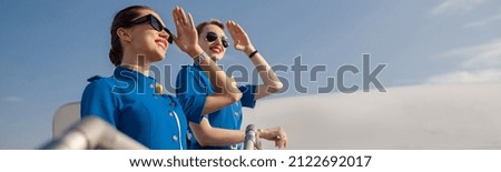 Portrait of two elegant air stewardesses in blue uniform and sunglasses covering eyes with hand and looking far away, standing together on airstair. Aircrew, occupation concept Royalty-Free Stock Photo #2122692017
