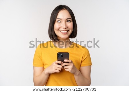 People and technology concept. Smiling asian girl using smartphone, texting on mobile phone, standing against white background