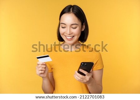 Online shopping. Smiling asian girl using credit card and mobile phone app, paying contactless, order on smartphone application, standing over yellow background