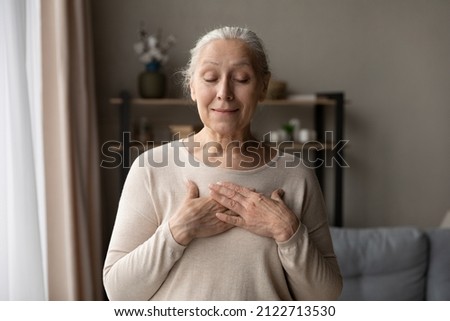 Happy grateful older 70s lady posing at home with closed eyes, applying hands to chest, heart, smiling, expressing gratitude, hope, love, appreciation, making wish. Mature woman showing kindness sign Royalty-Free Stock Photo #2122713530