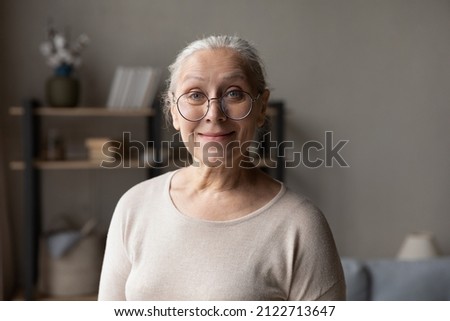 Smiling elderly grey haired lady in stylish glasses home head shot portrait. Happy senior retired woman holding video call, looking at camera, posing indoors with living room interior in background