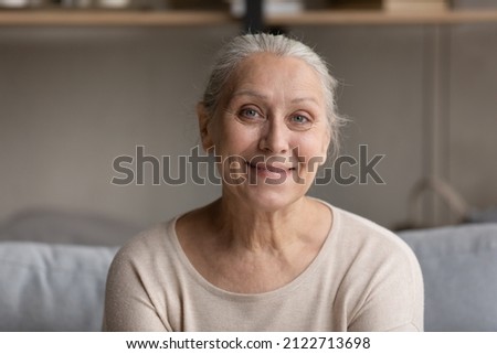 Smiling grey haired elder 70s lady head shot portrait. Happy senior woman sitting on sofa, posing at home, looking at camera, webcam during video call. Screen view. Elderly age concept