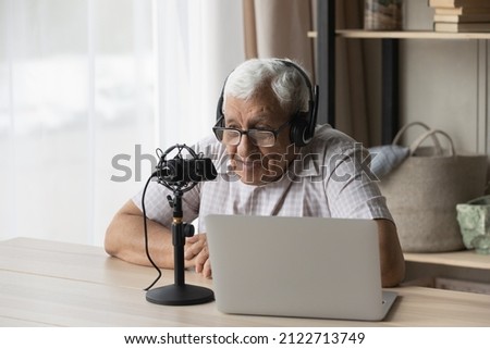 Elder radio host man in headphones and glasses speaking at professional microphone at laptop, holding program on air, broadcasting, recording audio book, using sound studio equipment at home