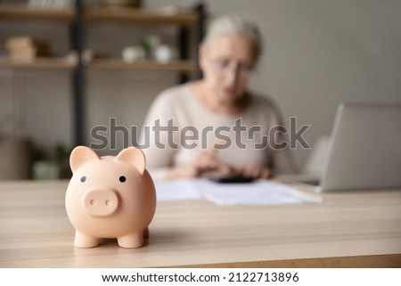 Toy pink piggy bank on work table of senior tenant, homeowner woman. Elderly lady using calculator, counting savings, taxes. Finance management, financial insurance, personal budget concept Royalty-Free Stock Photo #2122713896