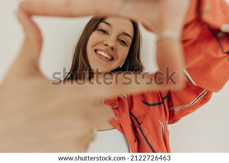 Photo of stylish young woman makes photo frame with her fingers on white wall background. Brown-haired woman smiles broadly with teeth wearing red shirt. Concept of invigorating mood.