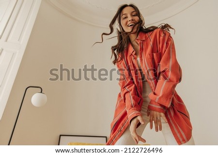 Portrait of happy young woman enjoys listening to music through headphones in bright room. Smiling beauty with wavy dark hair, in red shirt and white pants. Audio technology, gadgets and music concept Royalty-Free Stock Photo #2122726496