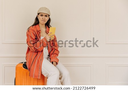 Caucasian teenager with disgruntled expression looks at camera and sits on suitcase indoors. Girl is holding modern smartphone in hands, dressed in shirt, pants. Emotion concept, facial expression.