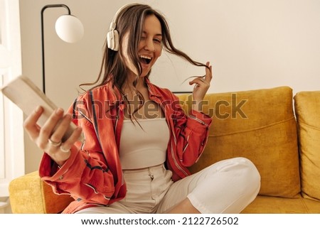 Fashionable european girl in good mood poses for photo while sitting on couch. Beauty in summer outfit relaxes while listening to favorite song through headphones. Lifestyle concept, playful mood.