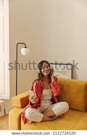 Portrait of young stylish woman sitting cross-legged on sofa at home with closed eyes. Girl wearing casual clothes, smiling enjoying weekend. Brown-haired listens to music through headphones. Royalty-Free Stock Photo #2122726520