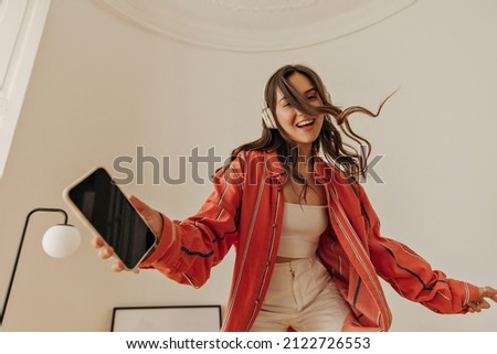 Energetic young model with wireless headphones is having lot of fun in closed white room. Dark-haired girl wear casual clothes holds phone in her hand and smiles cutely at camera. Royalty-Free Stock Photo #2122726553