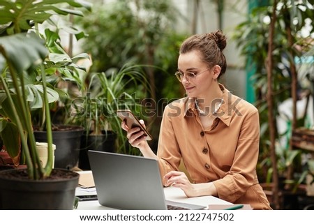 Young smiling businesswoman in brown blouse scrolling in mobile phone while communicating or looking through online information Royalty-Free Stock Photo #2122747502