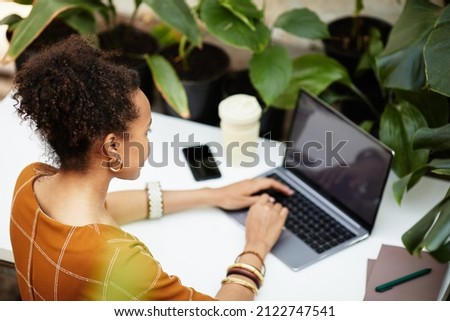 Young African American woman looking at laptop screen while typing on keyboard by desk and making order in online shop