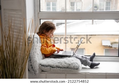 Little toddler girl sits on wide windowsill, plays games on laptop, listens to music on headphones. Child in an orange hoodie, gray pants at the window of the house. Distance learning for preschoolers