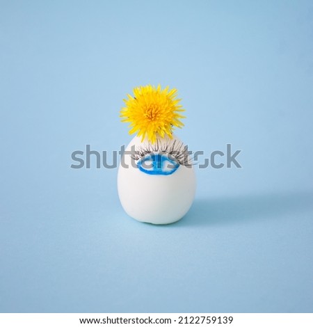 Creative white Easter egg with cute painted eye, eyelashes and yellow dandelion flower on pastel blue background. Happy Easter concept. Side view, interesting concept.