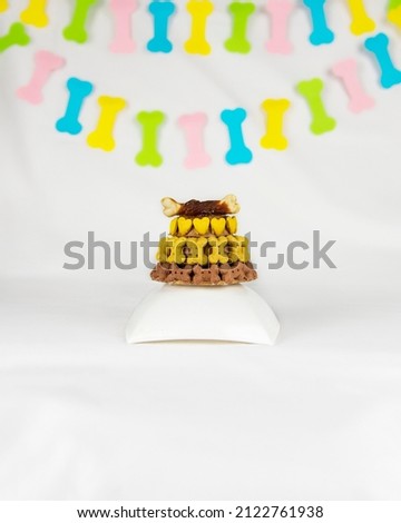 A three-tiered cake is for a dog stands on a white stand on a white background with a colored garland in the form of bones.Decorated with small yellow bones and hearts, with a bigger bone at the top 