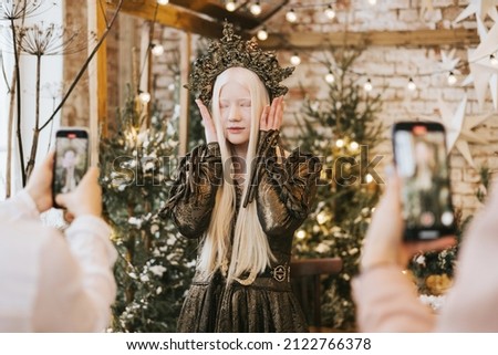 young woman with blue eyes and long white hair in beautiful green dress and crown stands in loft room decorated with wooden greenhouse and Christmas trees with twinkle lights, diverse people
