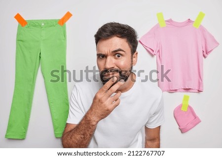 Serious man keeps hand on chin looks attentively at camera looks scrupulous raises eyebrows and examines something wears casual t shirt isolated over white background clothes plastered behind