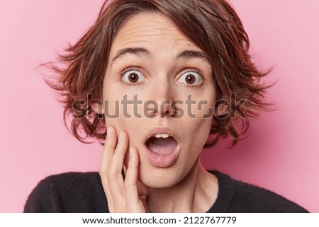 Close up shot of amazed scared woman stares at horrible thing holds breath keeps mouth widely opened keeps hand on cheek cannot believe her eyes feels frightened poses indoor. Human reactions Royalty-Free Stock Photo #2122767779
