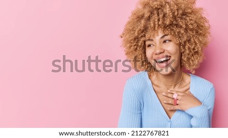 Positive curly haired young woman looks away with joyful expression smiles broadly expresses happy emotions wears casual blue jumper isolated over pink background copy space for your promotion Royalty-Free Stock Photo #2122767821