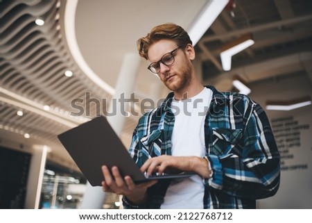 Bottom view of caucasian guy watching something on laptop computer in office space. Concept of flexible workplace. Idea of freelance and remote work. Young focused bearded successful man in glasses