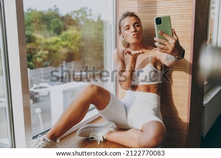 Young skinny sportswoman woman 20s in white sportswear earphones listen music do selfie shot on mobile cell phone blow air kiss sit on windowsill of gym rest indoors People urban lifestyle concept.