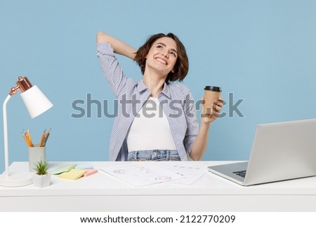 Young dreamful secretary employee business woman in casual shirt sit work at white office desk with pc laptop hold cup of coffee hold hand behind neck head isolated on pastel blue background studio. Royalty-Free Stock Photo #2122770209