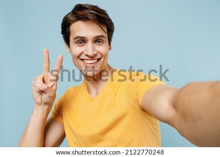 Close up young smiling happy satisfied fun cool man 20s wearing yellow t-shirt doing selfie shot pov on mobile phone show v-sign gesture isolated on plain pastel light blue background studio portrait.