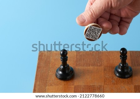 A man's hand with a white pawn over the chessboard. A QR code is attached to the base of the white pawn. There are two black pawns on the chessboard.                               