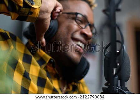 Close-up of an African podcaster blogger smiling while broadcasting his live audio podcast in the studio using a microphone and headphones. Male radio host with glasses, podcast host or interviewer