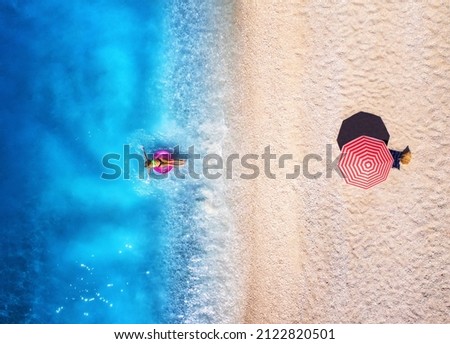 Aerial view of a woman swimming with pink swim ring in blue sea, sandy beach and red umbrella at sunset in summer. Tropical landscape with girl, clear water, waves. Top view. Lefkada island, Greece Royalty-Free Stock Photo #2122820501