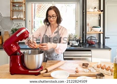 Woman wearing apron baking cookies in cozy kitchen. Housewife breaking eggs into a bowl of planetary mixer. Homemade pastries or cake. Royalty-Free Stock Photo #2122824509
