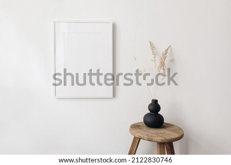 Portrait empty picture frame mockup in sunlight. Dry festuca, bunny tail grass in modern black organic shaped vase. Wooden stool, chair. White wall background. Scandinavian interior, home design. Art.