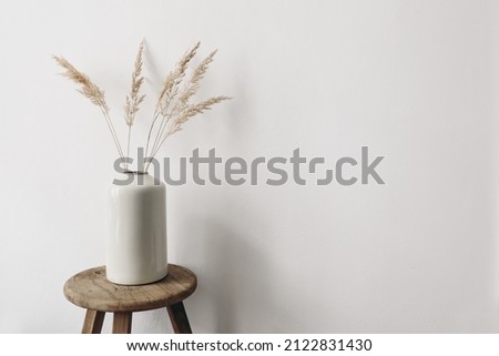 Modern summer, fall still life photo. Grey ceramic vase with dry festuca grass on old wooden stool. White wall background. Empty copy space. Elegant lifestyle decorative scene. Trendy interior decor. Royalty-Free Stock Photo #2122831430