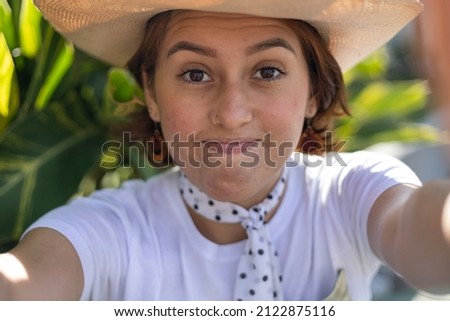 Young Latin American woman (22) with cowboy hat is sitting in the park smiling making a self-portrait with her cell phone. Concept Technology.