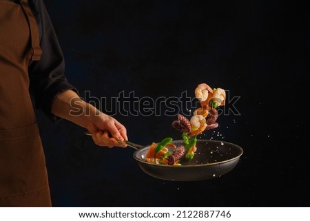 Cooking seafood - shrimp, octopus with vegetables in a frying pan on a black background by a professional chef. Frozen in-flight food. Sea food. Vegetarian, diet food. Banner. Royalty-Free Stock Photo #2122887746