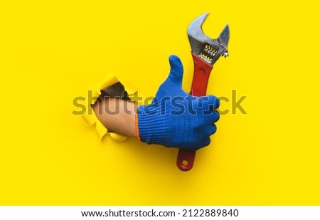 The right hand in a blue knitted glove holds a red wrench and gives a thumbs up (like). Torn hole in yellow paper. The concept of a worker, a labor migrant, a master of his craft. Copy space. Royalty-Free Stock Photo #2122889840