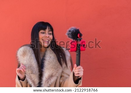 Smiling brunette girl recording a video blog with her mobile phone and microphone, wearing a winter jacket for social networks. Caucasian pretty a red background