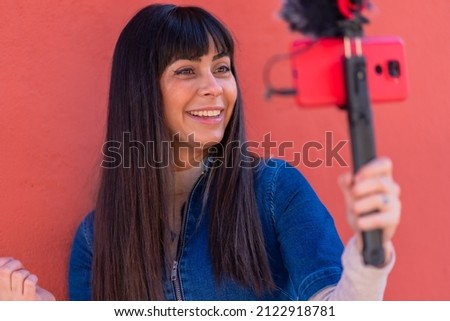 Brunette girl smiling recording a video blog with the mobile phone and microphone, wearing a blue denim suit for social networks. Caucasian pretty a red background