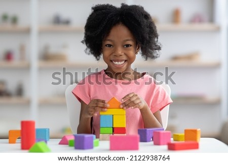 Happy little african american girl with bushy hair playing with colorful wooden blocks and smiling at camera, enjoying table games while visiting psychologist or playing alone at home, closeup Royalty-Free Stock Photo #2122938764